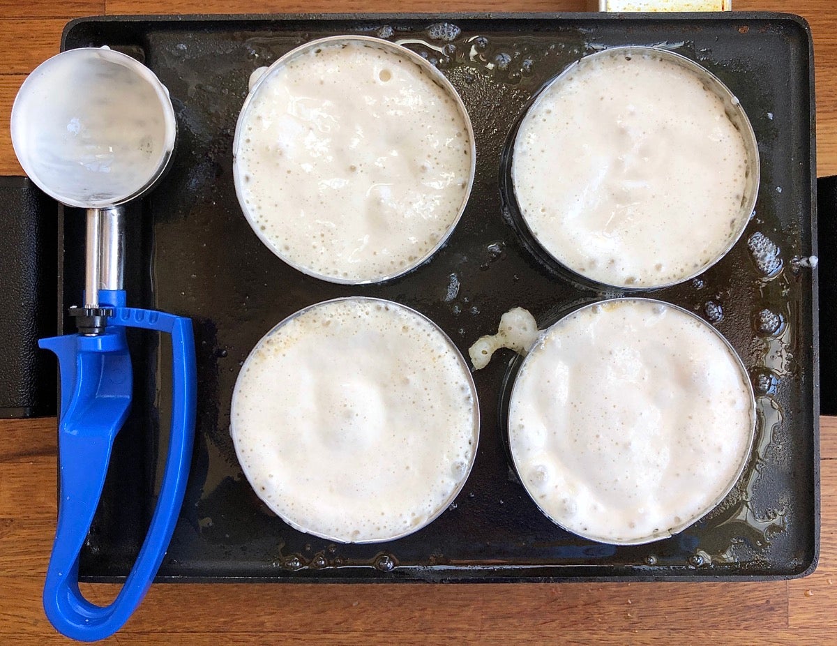 Four muffin rings on a hot griddle filled with cooking sourdough crumpet batter.